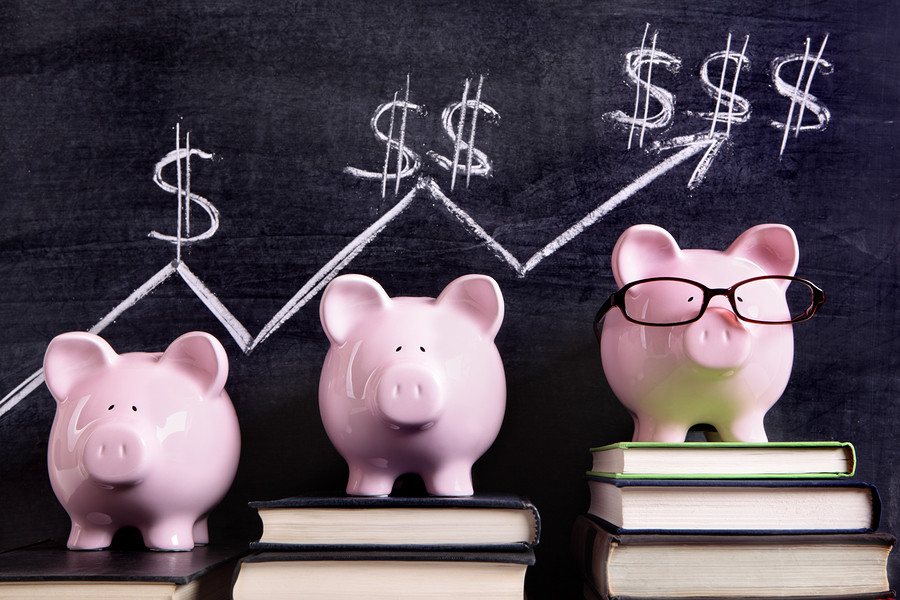 Three pink piggy banks standing on books next to a blackboard