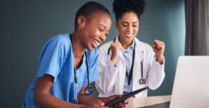 Two front-line healthcare workers celebrate while reviewing pay or performance updates on a digital device, representing how modern compensation management empowers and supports front-line teams.