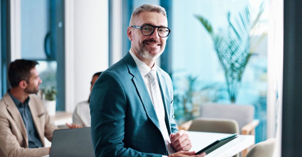A senior leader in a teal suit stands with a tablet in a modern private equity office, while colleagues meet in the background to discuss firm growth and talent strategy.