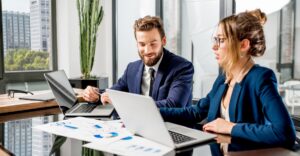 Two financial services professionals review compensation data together on laptops, with printed charts visible on the conference table.