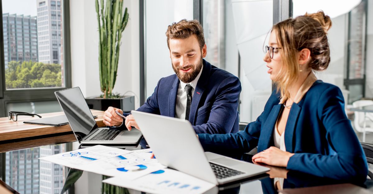 Two financial services professionals review compensation data together on laptops, with printed charts visible on the conference table.