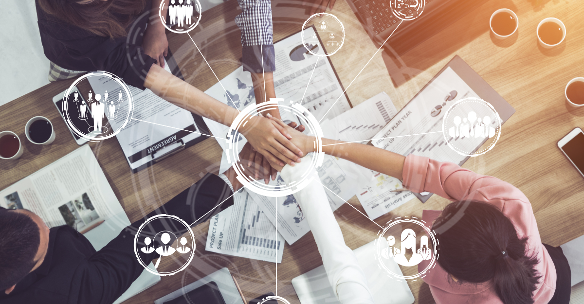 Overhead view of business team shaking hands over a wooden table with documents and charts, surrounded by holographic icons representing people, teamwork, and organizational structure connected by lines.